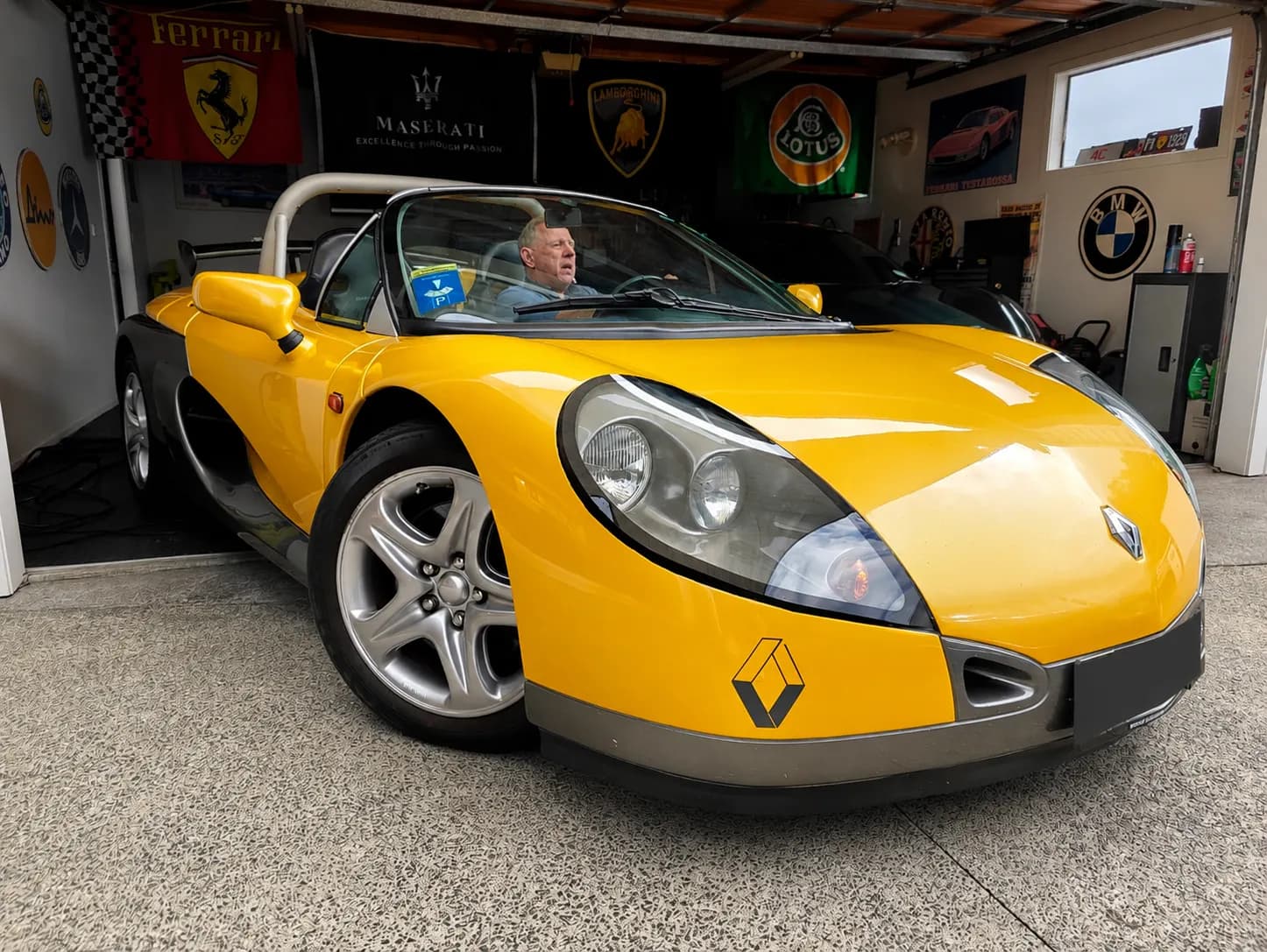 Yellow Renault Sport Spider parked inside a private enthusiast garage with Ferrari, Maserati, Lamborghini, Lotus, and BMW banners displayed in the background.